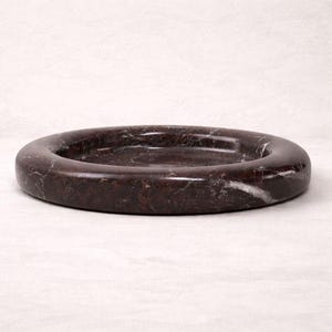 May include: A dark brown marble ashtray with a circular design. The ashtray has a smooth, polished surface with white veining. The ashtray is a decorative item for the home.