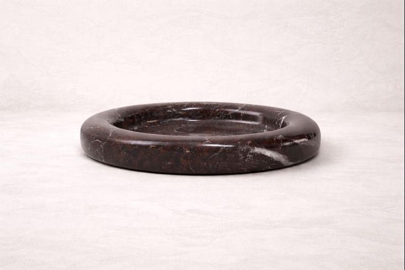 May include: A dark brown marble ashtray with a circular design. The ashtray has a smooth, polished surface with white veining. The ashtray is a decorative item for the home.