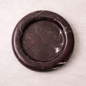 May include: A dark brown marble coaster with white veining. The coaster has a circular shape with a raised edge. This decorative item is suitable for holding beverages and protecting surfaces.