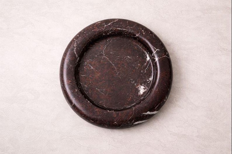 May include: A dark brown marble coaster with white veining. The coaster has a circular shape with a raised edge. This decorative item is suitable for holding beverages and protecting surfaces.