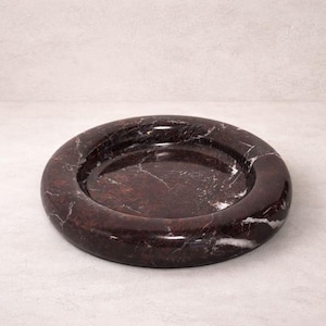 May include: A round, dark brown marble dish with white veining. The dish has a raised, rounded rim and a shallow interior. The marble has a polished finish, and the dish is set against a neutral background.