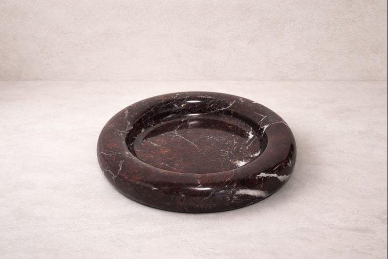 May include: A round, dark brown marble dish with white veining. The dish has a raised, rounded rim and a shallow interior. The marble has a polished finish, and the dish is set against a neutral background.