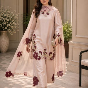 May include: A light pink long-sleeved tunic and wide-leg trousers set featuring dark maroon floral embroidery along the hemlines and neckline. A matching sheer pink dupatta with maroon floral appliqu&eacute;s drapes over the shoulders. Text reads Rangsaari.