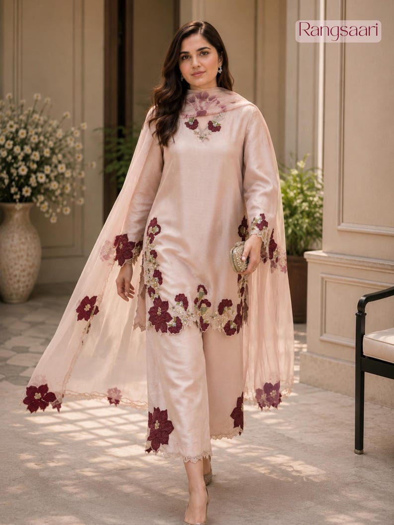 May include: A light pink long-sleeved tunic and wide-leg trousers set featuring dark maroon floral embroidery along the hemlines and neckline. A matching sheer pink dupatta with maroon floral appliqu&eacute;s drapes over the shoulders. Text reads Rangsaari.