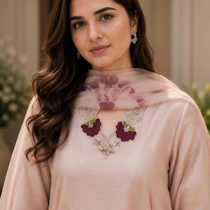 May include: A light peach-coloured tunic featuring a keyhole neckline adorned with dark maroon floral embroidery and silver accents. A matching sheer peach dupatta with a subtle purple floral pattern is draped over the shoulders. The brand name Rangsaari is visible in the top right corner.