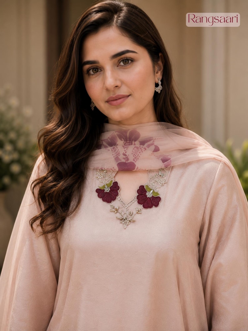 May include: A light peach-coloured tunic featuring a keyhole neckline adorned with dark maroon floral embroidery and silver accents. A matching sheer peach dupatta with a subtle purple floral pattern is draped over the shoulders. The brand name Rangsaari is visible in the top right corner.