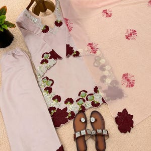 May include: A light pink three-piece outfit consisting of a long-sleeved tunic, wide-leg trousers, and a sheer dupatta. The tunic features dark red floral appliqu&eacute;s, silver sequins, and light green accents. A pair of brown sandals is included.