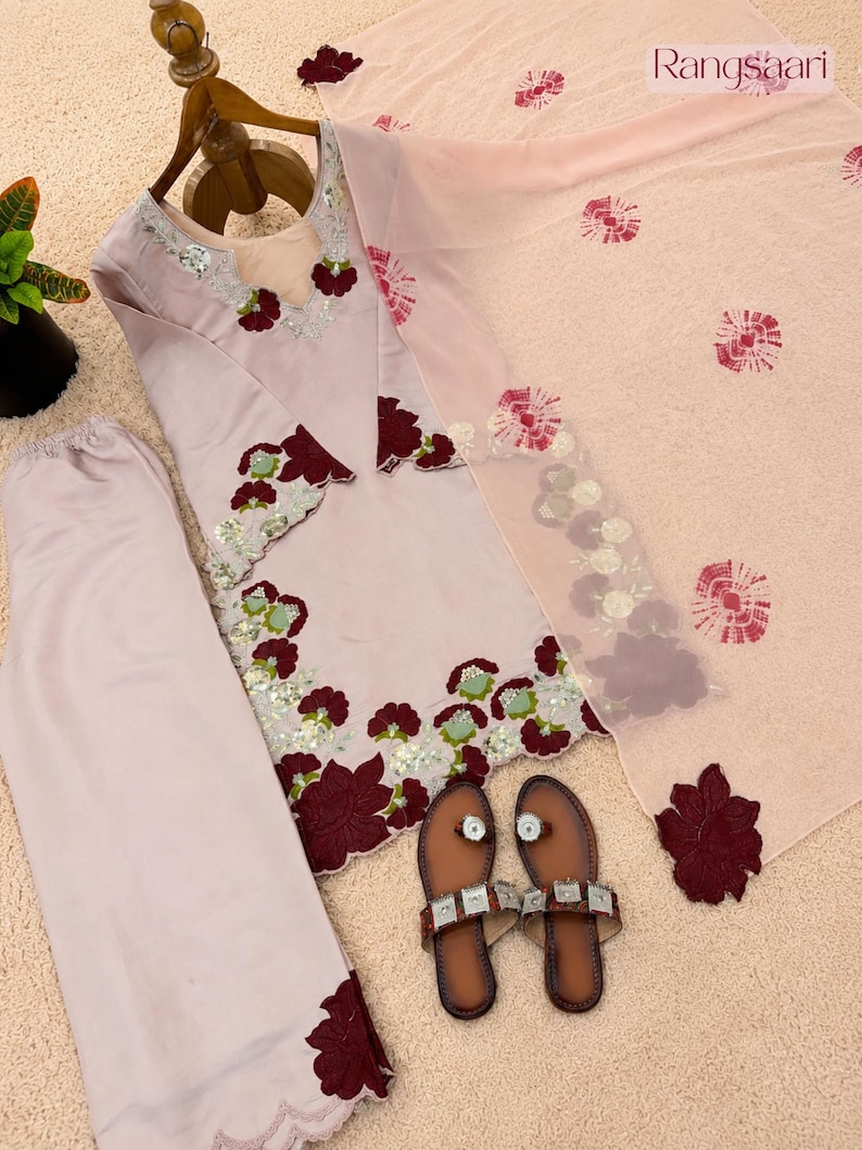 May include: A light pink three-piece outfit consisting of a long-sleeved tunic, wide-leg trousers, and a sheer dupatta. The tunic features dark red floral appliqu&eacute;s, silver sequins, and light green accents. A pair of brown sandals is included.