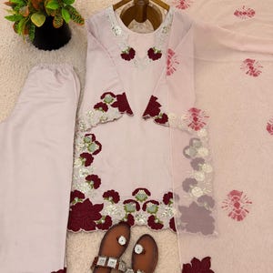 May include: A light pink three-piece outfit consisting of a long-sleeved tunic, matching trousers, and a dupatta. The tunic features dark red floral embroidery, silver sequins, and scalloped edges. The dupatta has a tie-dye pattern and the text Rangsaari.