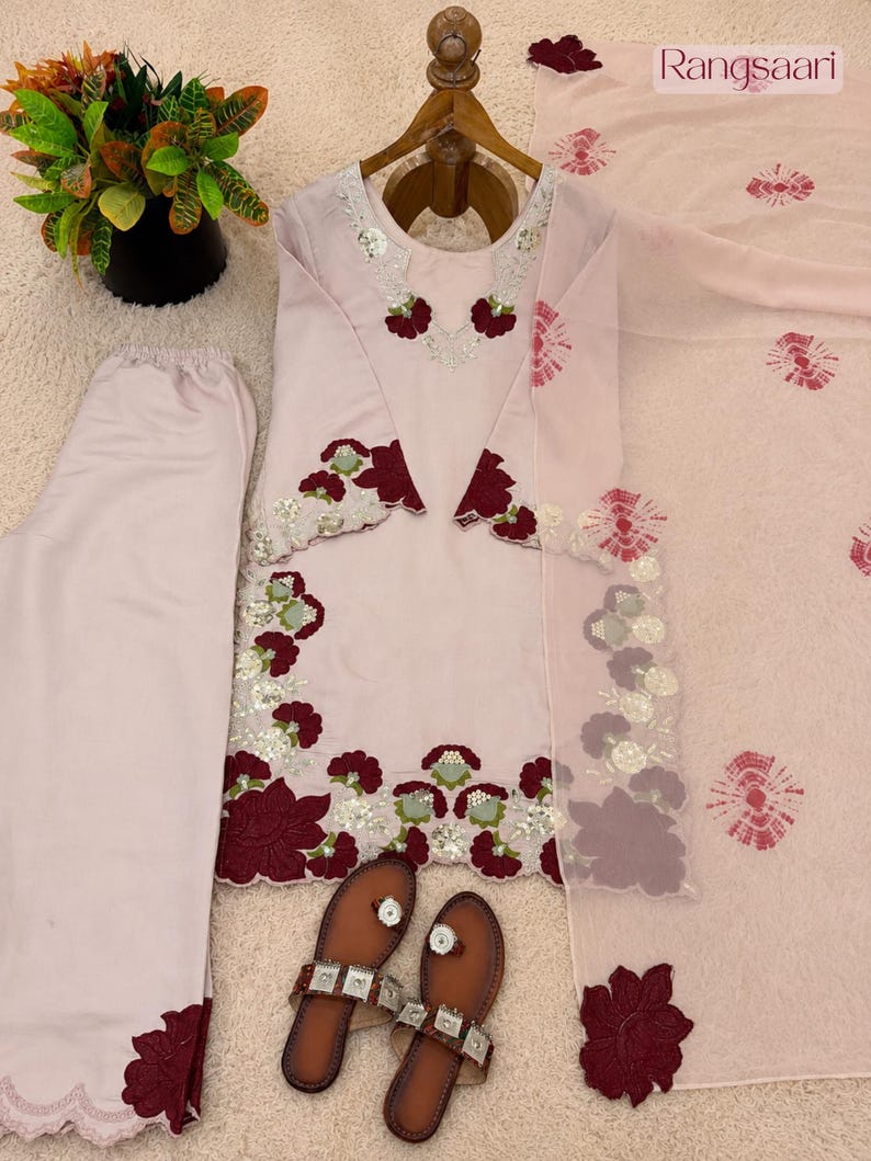 May include: A light pink three-piece outfit consisting of a long-sleeved tunic, matching trousers, and a dupatta. The tunic features dark red floral embroidery, silver sequins, and scalloped edges. The dupatta has a tie-dye pattern and the text Rangsaari.