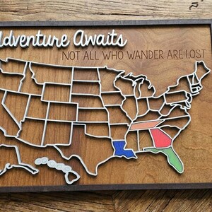 May include: Wooden map of the United States with state outlines, framed in dark brown. The map has a white outline and includes the text "Adventure Awaits" and "Not All Who Wander Are Lost". Some states are colored.