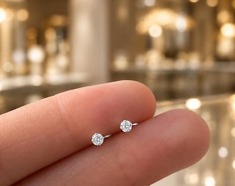 May include: Two small, round, clear stud earrings featuring a single brilliant-cut stone set in a silver-toned metal prong setting. The earrings are positioned on a finger, showcasing their compact size and reflective, sparkling appearance.