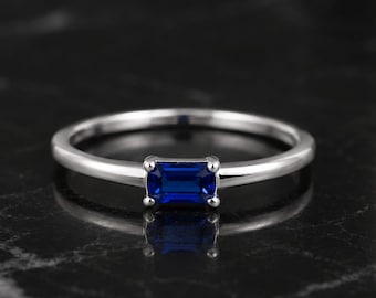 May include: A silver-toned ring featuring a single, rectangular, emerald-cut blue gemstone set in a four-prong setting. The band is smooth, polished, and circular, showcasing a minimalist design with a vibrant, deep blue center stone.