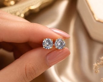 May include: A pair of round, brilliant-cut clear stud earrings set in gold-toned metal prongs. Each earring features a single faceted stone that reflects light, showcasing a classic circular design held together by a secure metal backing post.