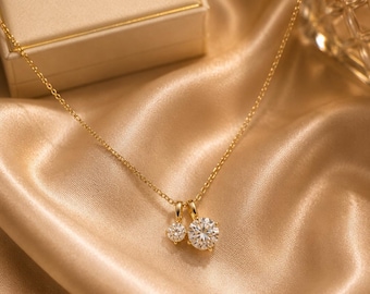 May include: A gold chain necklace featuring two round, clear, faceted stone pendants of different sizes. The necklace is positioned near a beige jewelry box with the gold-embossed text "D DAS N JEWELS" on the lid, set against a draped fabric.