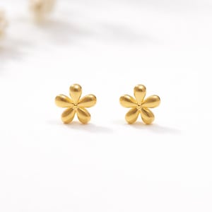 May include: A pair of small, gold-colored flower stud earrings. Each earring features five petals and a rounded center, set against a white background. The earrings are a delicate floral design.