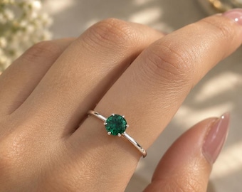 May include: A silver ring featuring a single round-cut green gemstone held in a six-prong setting. The band is thin and polished, showcasing a minimalist design that highlights the vibrant emerald-colored stone centered on the finger.