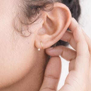 Puede incluir: Primer plano de una oreja adornada con dos pendientes dorados. Uno es un aro peque&ntilde;o y el otro es un peque&ntilde;o pendiente con una gema brillante. La mano toca suavemente la oreja, resaltando las joyas.