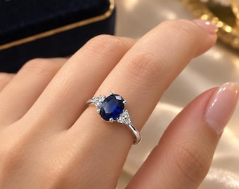 May include: A silver-toned ring featuring a central oval-cut deep blue gemstone flanked by three small clear stones on each side. The band is thin and polished, showcasing a classic prong setting that secures the vibrant blue center stone.