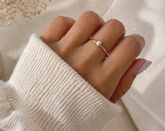 May include: A delicate gold ring with a small, round, white gemstone is worn on a finger. The ring is set against a backdrop of a cream-colored sweater and a soft, neutral-toned background. The ring's simple design highlights the gemstone.