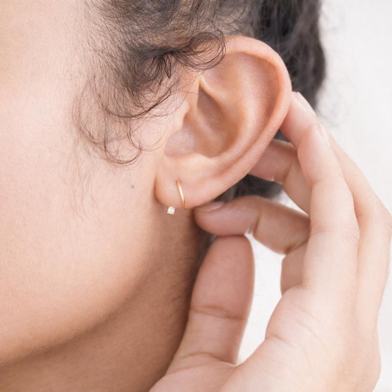 Puede incluir: Primer plano de una oreja adornada con dos pendientes dorados. Uno es un aro peque&ntilde;o y el otro es un peque&ntilde;o pendiente con una gema brillante. La mano toca suavemente la oreja, resaltando las joyas.