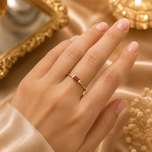 May include: A thin gold ring featuring a single rectangular baguette-cut red gemstone at the centre, flanked by two tiny clear accent stones on either side of the band. The metal band is polished and smooth, showcasing a minimalist design.