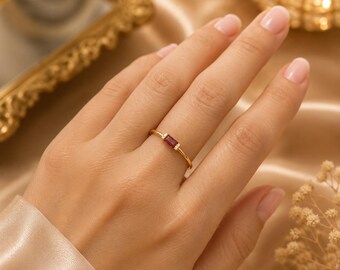 May include: A thin gold ring featuring a single rectangular baguette-cut red gemstone at the center, flanked by two tiny clear accent stones on either side of the band. The metal band is polished and smooth, showcasing a minimalist design.