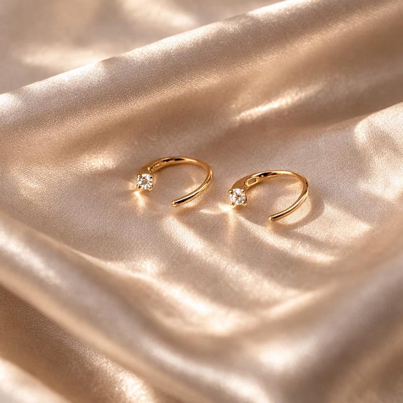Puede incluir: Un par de pendientes de aro dorados, cada uno con una peque&ntilde;a gema transparente. Los pendientes se presentan sobre un tejido sedoso de color marr&oacute;n claro. Un dise&ntilde;o sencillo y elegante. Pendientes de estilo cl&aacute;sico.