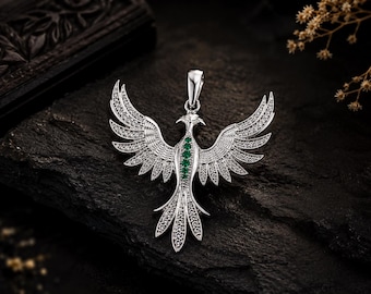 May include: A silver-toned pendant shaped like a bird with wings spread wide. The wings and tail are encrusted with numerous small, clear, sparkling stones. A vertical row of four small, vibrant green stones is set along the center of the bird's body.