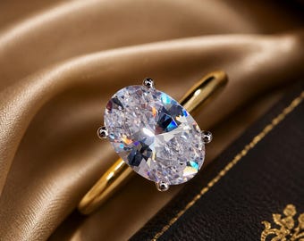 May include: A gold ring featuring a large, clear, oval-cut gemstone held in place by four silver-toned prongs. The band is smooth and polished, showcasing a reflective metallic finish that complements the brilliant, faceted stone at the center.