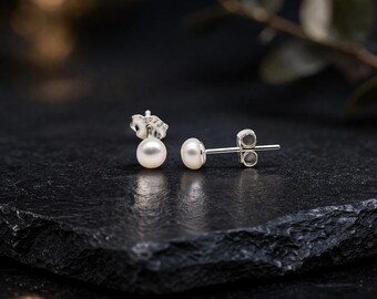 May include: A pair of pearl stud earrings. Each earring features a small, round, white pearl set on a silver post with a butterfly backing. The earrings are displayed on a dark, textured surface, with a blurred background.