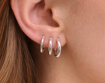 May include: Three silver hoop earrings of varying sizes are worn in the ear. The earrings are shiny and reflect light. The ear is visible, with the skin tone appearing fair. The hair is a light brown color.