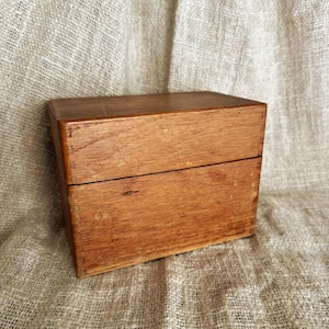May include: A rectangular wooden box with a hinged lid. The box is made of light brown wood and has visible wood grain. The box is sitting on a textured, light brown surface.