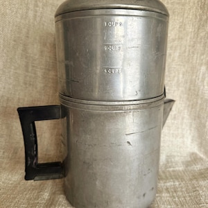 Può includere: Una caffettiera a percolazione vintage in alluminio con manico e coperchio neri. La caffettiera ha misure per 4, 6 e 8 tazze. Un classico elettrodomestico da cucina.