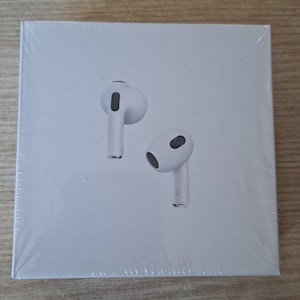 May include: A square white product box featuring a graphic illustration of two white wireless earbuds on the front. The packaging is sealed in clear plastic wrap. One earbud is positioned above the other, and the lower earbud has an R printed on it.