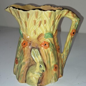 Puede incluir: Jarra de cerámica decorativa con un diseño inspirado en la naturaleza. La jarra tiene un fondo amarillo con un patrón de espigas de trigo, amapolas naranjas y un conejo verde claro. El asa tiene forma de rama de árbol.