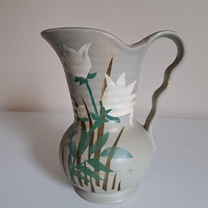 Può includere: Brocca in ceramica grigia con manico e beccuccio curvi. La brocca è decorata con motivi floreali bianchi, foglie verdi e steli marroni. Ha una base bulbosa e una parte superiore leggermente svasata.
