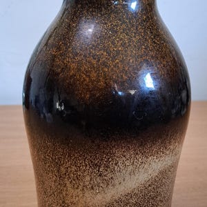 May include: A brown ceramic vase with a speckled, textured finish. The vase has a wide base that tapers to a narrow neck and flared rim. The color transitions from dark brown to tan. The vase is suitable for home decor.