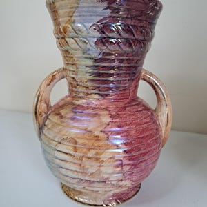 May include: A decorative ceramic vase with two handles, featuring a textured, ribbed design. The vase has a wide opening and a bulbous body. The color palette includes shades of pink, purple, beige, and blue, with iridescent highlights. The vase is approximately 10 inches tall.