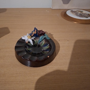 May include: A round, dark brown ceramic ashtray featuring a glossy, multi-colored glazed center. The rim is decorated with a sculpted white edelweiss flower, a blue leaf, and a small dark red floral accent, creating a textured, decorative edge.