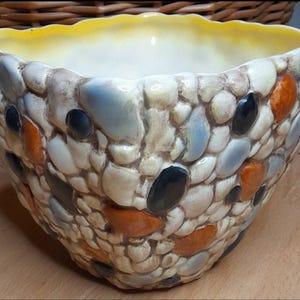 May include: A decorative bowl with a pebble design. The bowl's exterior is covered in a mosaic of rounded, multi-colored stones, including white, blue, orange, and black. The interior is a light yellow color.