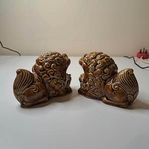 May include: A pair of brown ceramic Foo Dog statues. Each statue is in a seated position with detailed fur and mane. The statues have a glossy finish and are displayed on a white surface.