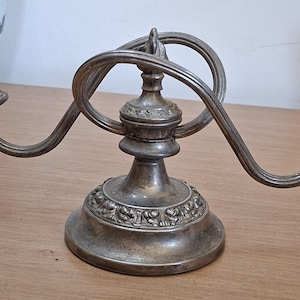 May include: A silver-toned metal candelabra featuring two candle holders extending from a central base. The base and candle cups are decorated with embossed floral patterns, and the central stem is topped with two interlocking circular rings.