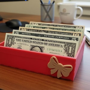 May include: A red rectangular container with a beige bow detail, filled with U.S. dollar bills. The bills are of various denominations, including one, five, and ten-dollar bills. The text "MOTION PICTURE PURPOSES" is visible on some bills.