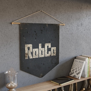 May include: A dark gray banner with the text "RobCo" in a distressed white font. The banner is hanging from a wooden dowel with a string. The banner is mounted on a gray wall.