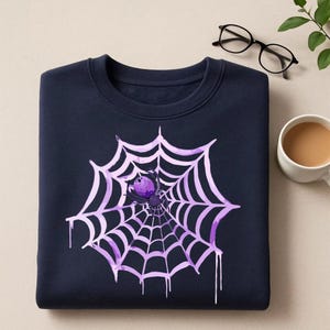 May include: Navy blue sweatshirt featuring a large, light purple spider web design with a purple spider at the center. The web has a dripping effect. A casual, comfortable top for Halloween or spooky season.