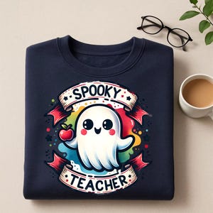 May include: Navy blue sweatshirt featuring a cartoon ghost holding a red apple. The ghost is surrounded by a rainbow-colored design and the words "Spooky Teacher" are written on a banner. A fun and festive design for Halloween.
