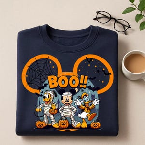 May include: Navy blue sweatshirt featuring a Halloween-themed graphic. The design includes Mickey Mouse, Pluto, and Donald Duck dressed as mummies, with pumpkins and the word "BOO!!" in orange. The sweatshirt has a large orange Mickey Mouse ear outline.