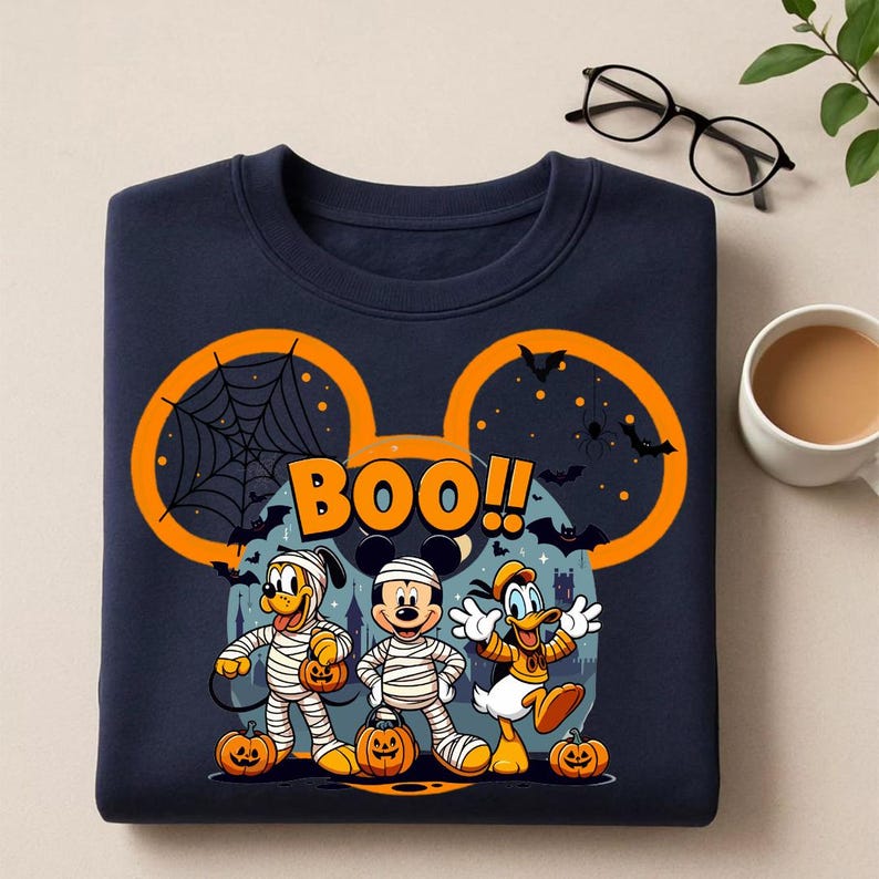 May include: Navy blue sweatshirt featuring a Halloween-themed graphic. The design includes Mickey Mouse, Pluto, and Donald Duck dressed as mummies, with pumpkins and the word "BOO!!" in orange. The sweatshirt has a large orange Mickey Mouse ear outline.