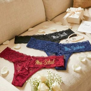 Könnte beinhalten: Vier Spitzen-Tanga-Höschen in Schwarz, Marineblau und Burgunderrot, jeweils mit einem Namen in Gold- oder Weißfaden bestickt. Ein Strauß weißer Rosen und eine kleine Geschenkbox sind ebenfalls vorhanden.